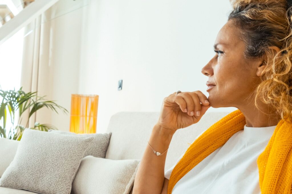 Mulher morena de aprox. 55 anos sentada no sofá, de perfil, com a mão no queixo