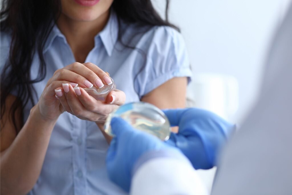 Mulher caucasiana de cabelos pretos segura uma prótese de silicone enquanto um profissional de saúde, à sua frente, segura outra prótese, ilustrando opções para cirurgia mamária.